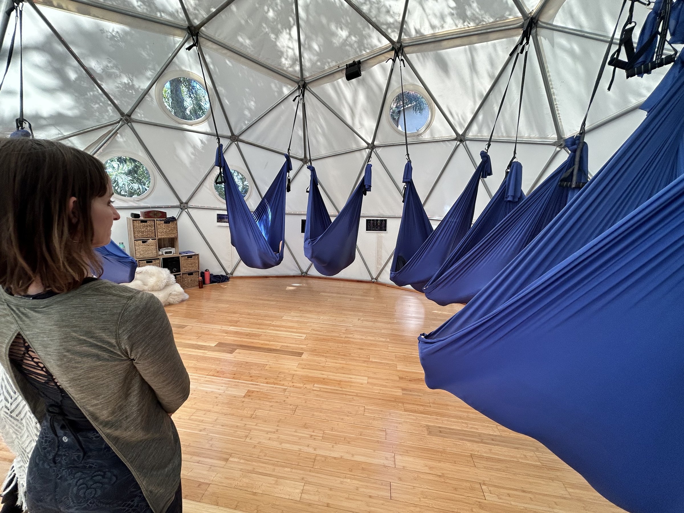 A group lying together in the hanging swings inside the Aum Dome, each person held in a swing of their own, while Maura stands at the edge of the room in side profile, teaching.