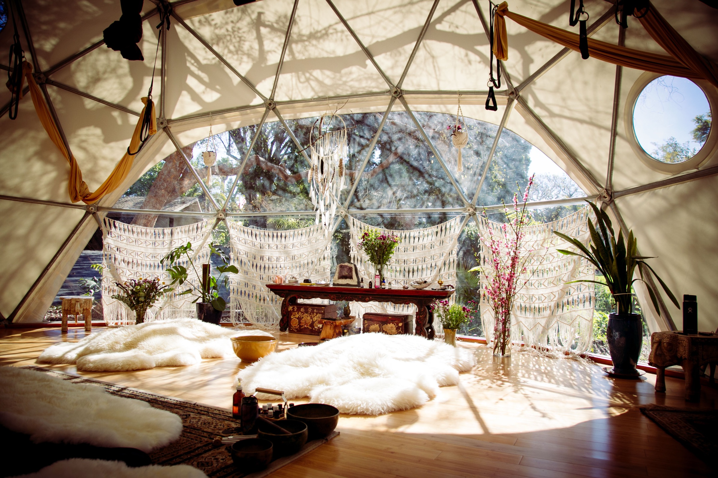 The interior of the Aum Dome at golden hour, cream geodesic panels above a bamboo floor, gold silks and sheepskins arranged on the ground