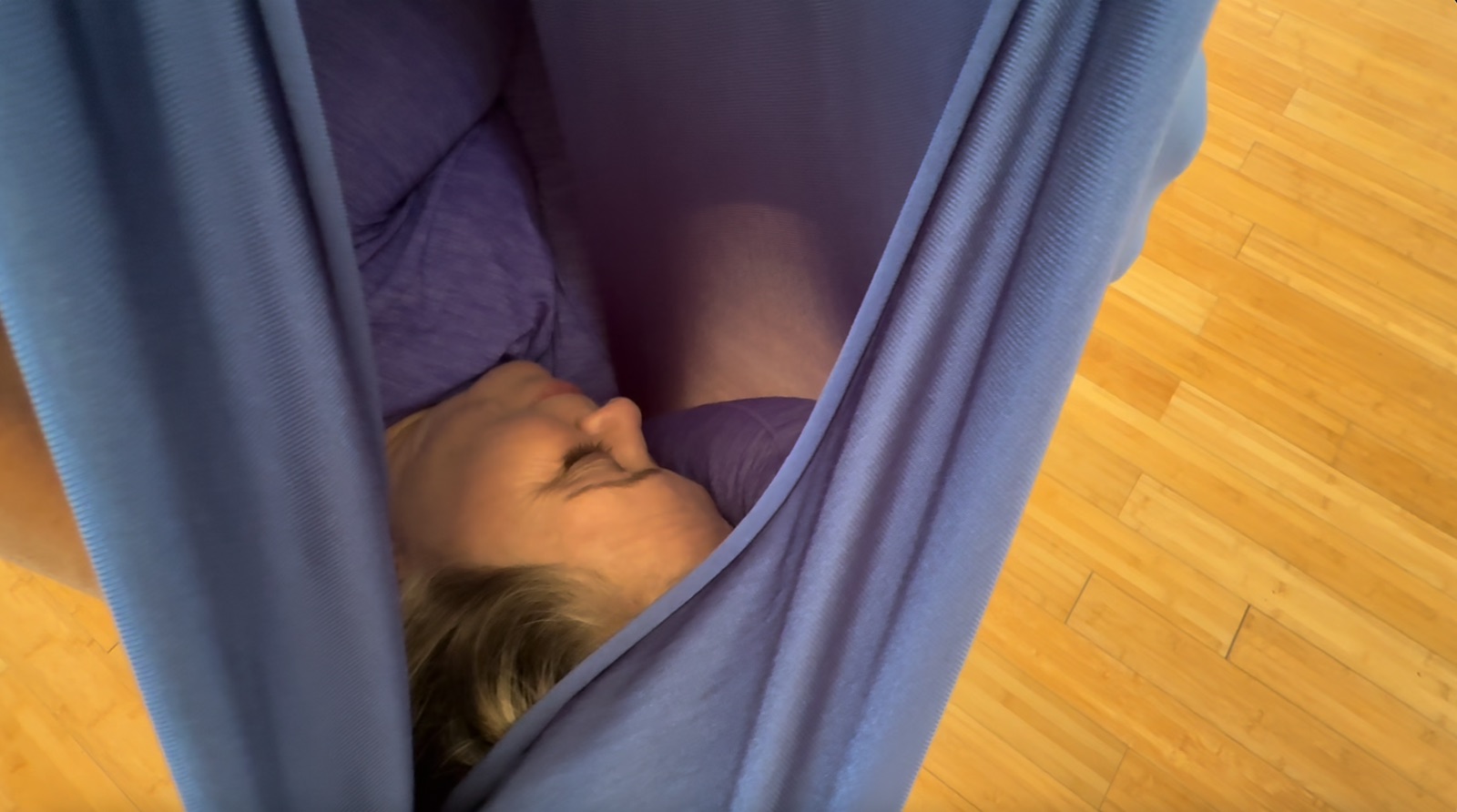 A client lying with their eyes closed inside the folded fabric of the swing, held entirely by the cloth.