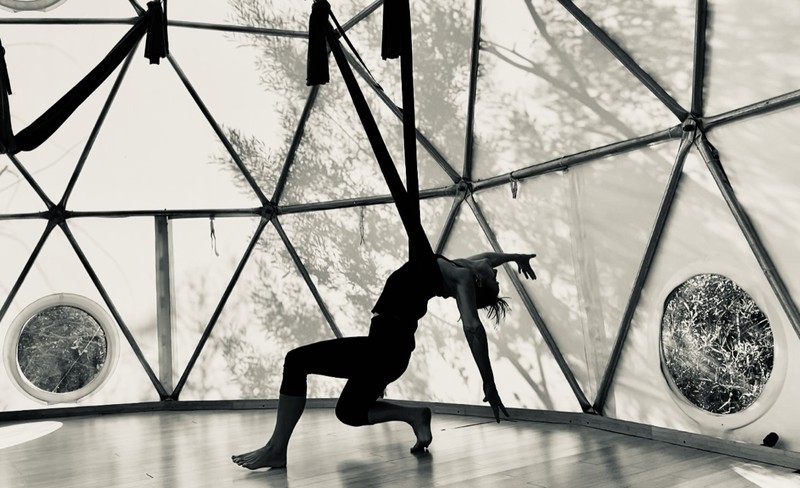 A black and white frame of a deep lunge inside the dome, the practitioner silhouetted against the geodesic frame with the swings hanging behind